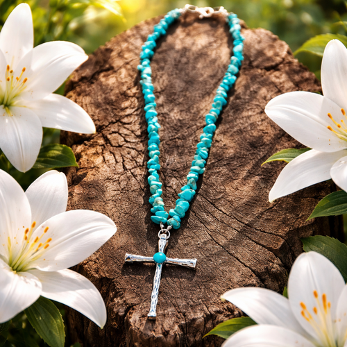 Turquoise beaded necklace with a silver cross pendant on a wooden stump surrounded by white lilies.